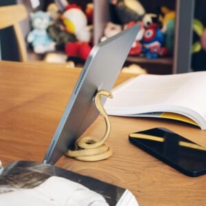Stylish cobra-shaped metal stand holding a tablet on a wooden desk with plush toys in background.