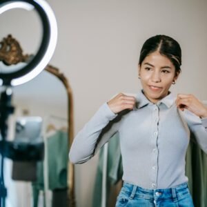 A young woman adjusting her blouse in front of a mirror, preparing for a fashion vlog indoors.