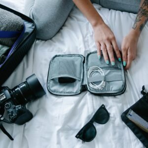 Woman with tattoos packs electronics, camera, and sunglasses in a suitcase on a bed.